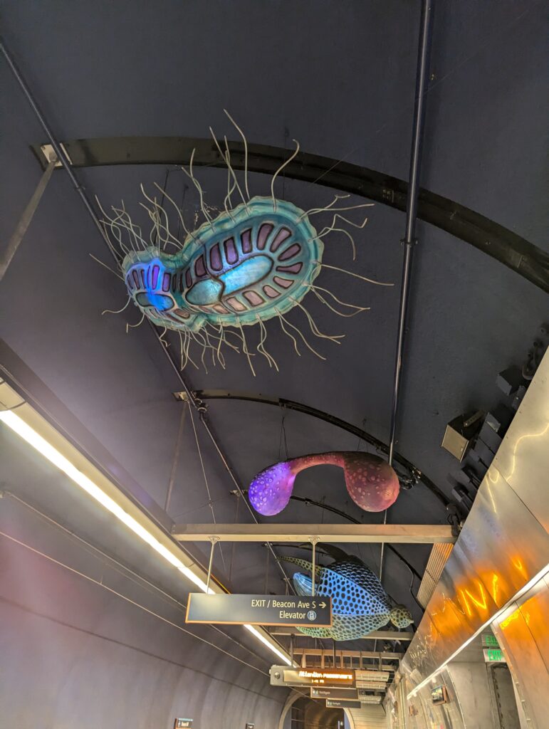 A scuplture that looks like a cross between an amoeba and a jellyfish, lit from inside, hanging from the ceiling of a light rail station. Two other sculptures are visible in the background.